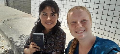 Alyssa and Sister Zurita on with the Book of Mormon sitting on the curb